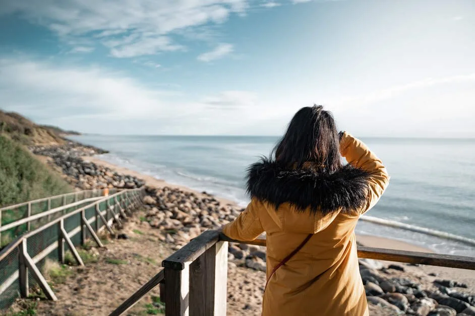 A woman in a yellow coat with a black fur-lined hood standing on a wooden platform overlooking the beach and ocean, shielding her eyes from the sun.