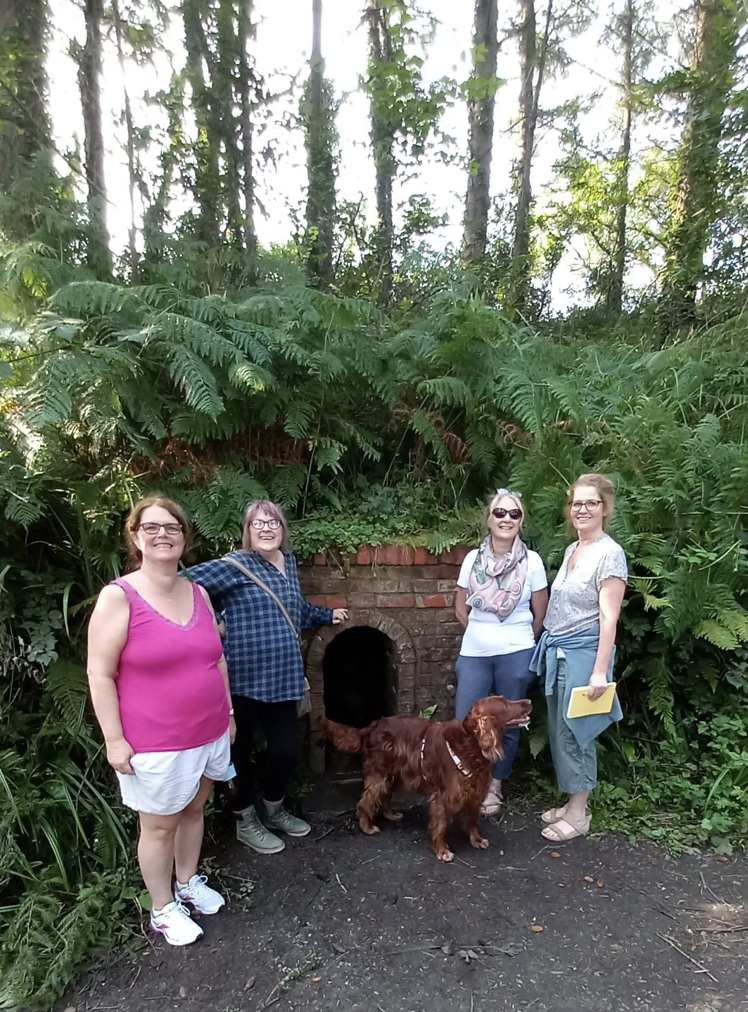 Four women and a dog standing in front of a brick oven built into a small hill covered with lush green plants and trees.
