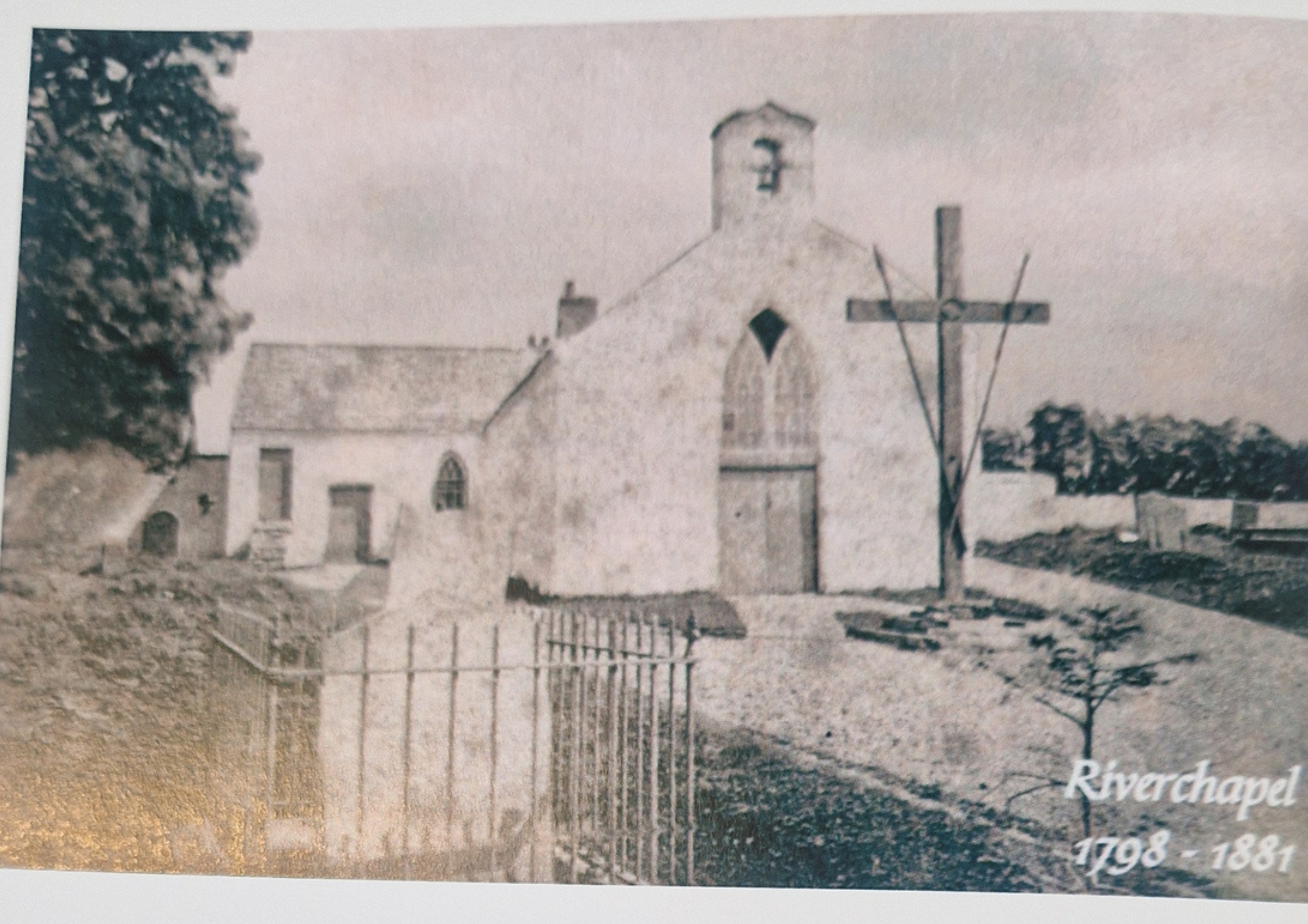Black and white photograph of a historic church building with a steeple, a cross, and a tree in the foreground. The image has the text 'Riverschapel 1908-1981' in the bottom right corner.