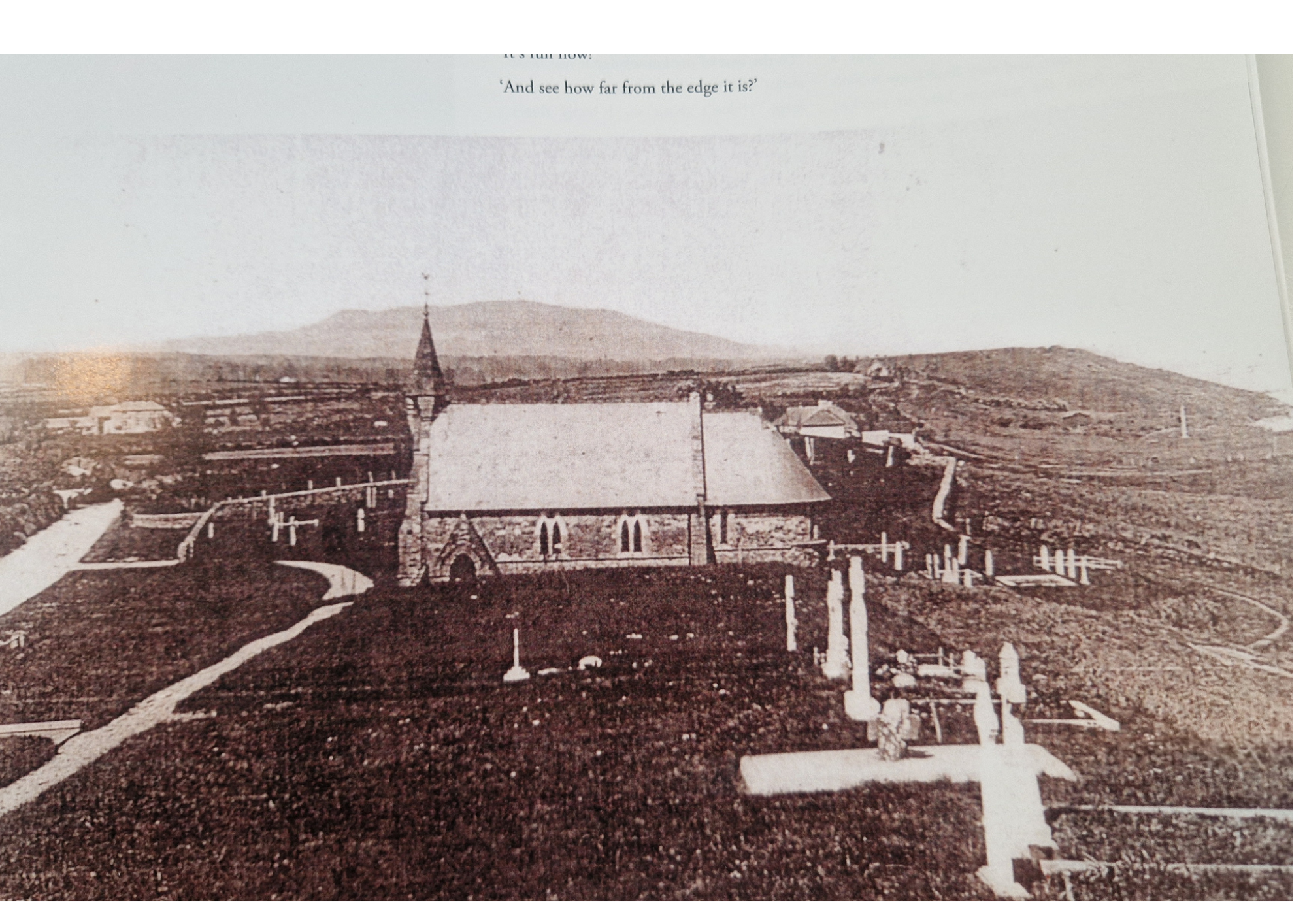 Old black-and-white photo of a church with a steeple, surrounded by a manicured yard with white posts and pathways, with a rural landscape and hills in the background.