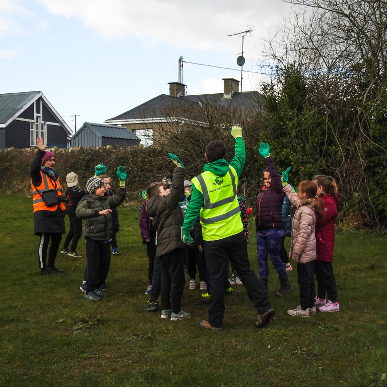 Group of children and adults outdoors on grassy field, with children raising hands and wearing jackets and gloves, assisted by adults in high-visibility vests.