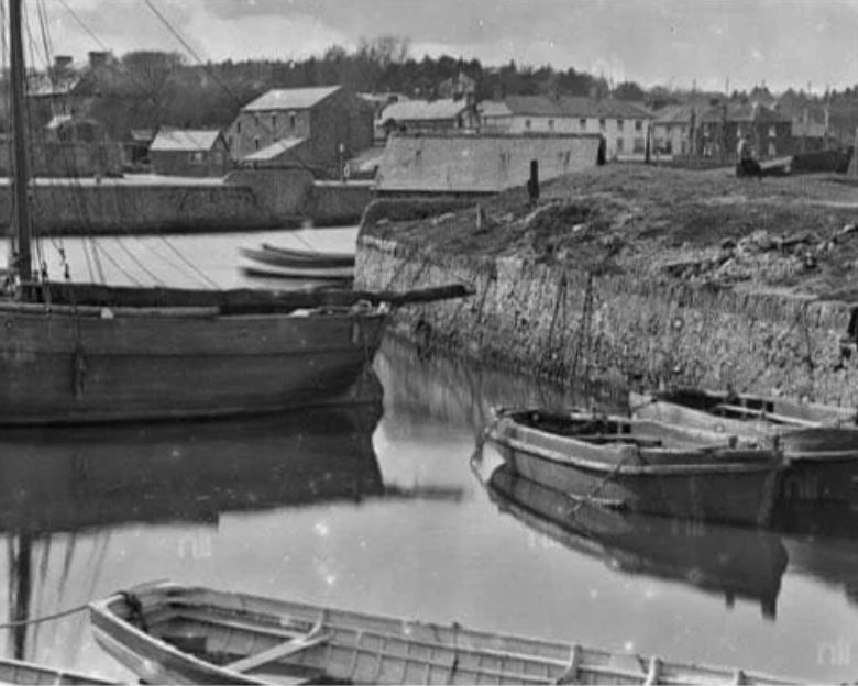 Black and white photo of a small harbor with boats docked, and buildings in the background.