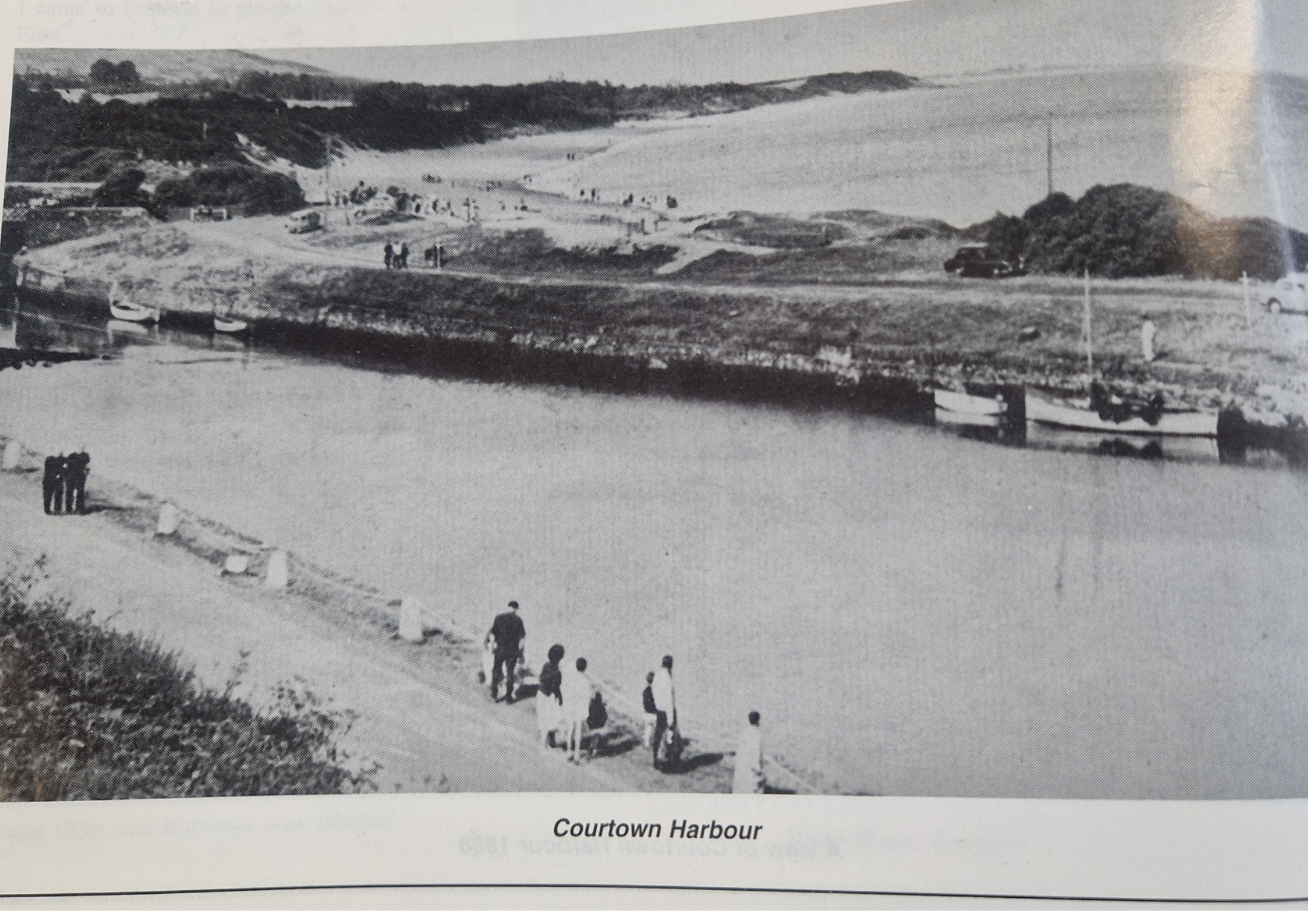 Black and white photograph of Courtown Harbour with people walking along a path by a body of water, boats docked, trees, and hills in the background.
