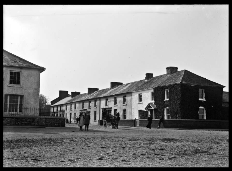 Black-and-white historic photograph of a row of houses with people and a horse-drawn carriage on the street in front.