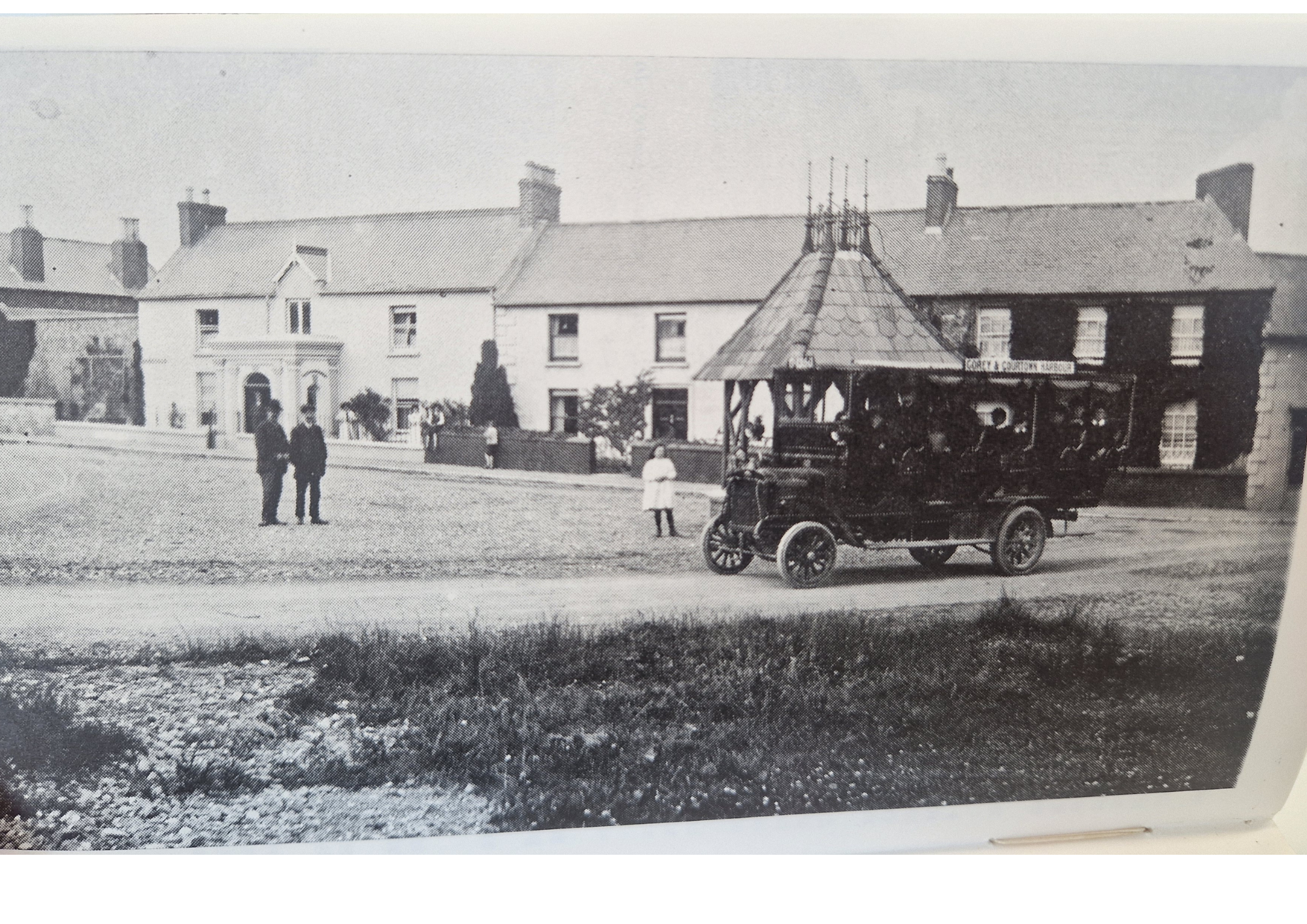 A black and white photograph of a street scene with several buildings, including one labeled "Kerry & Coulter Garage," and a vintage car parked in front. There are a few people walking or standing on the sidewalk.