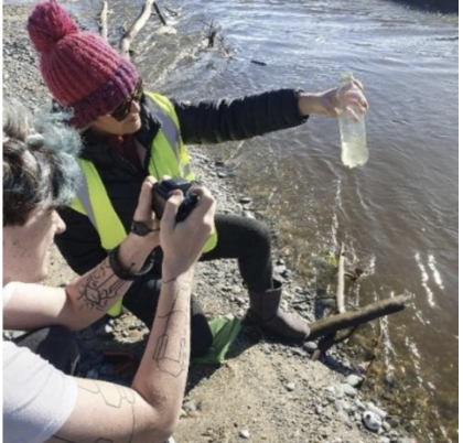 A woman with a red knit beanie and sunglasses taking a photo of a shallow river with a smartphone while a person with tattoos holds a clear plastic bottle over the water.