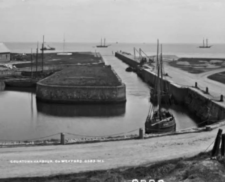 Black and white photo of a harbor with boats, a harbor wall, a narrow channel, and a body of water in the background.