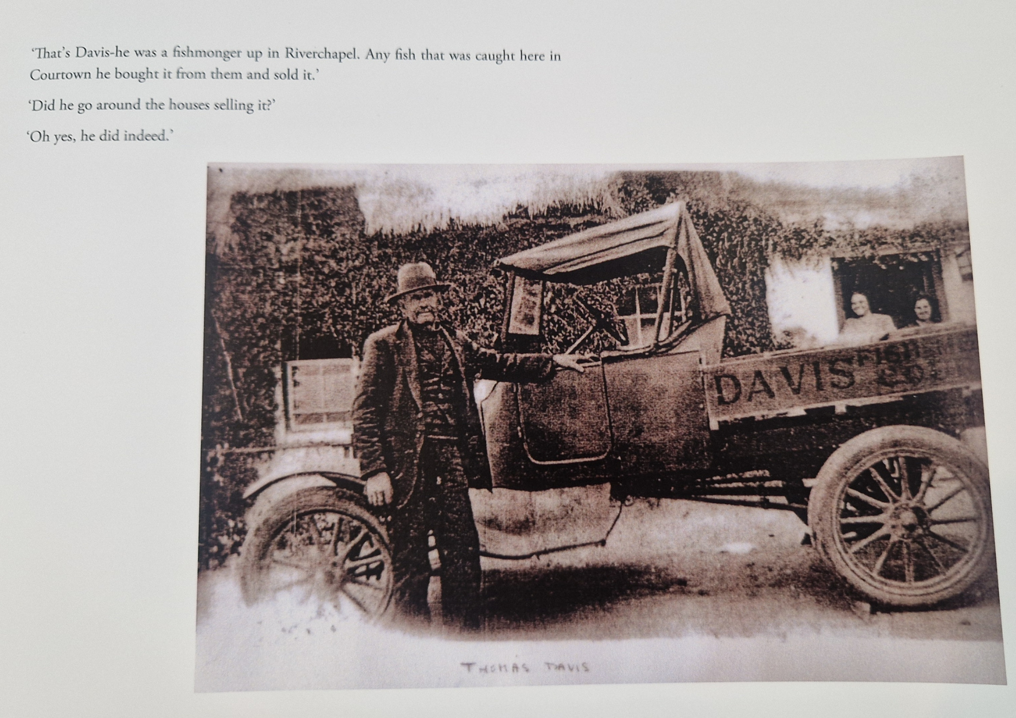 Old black and white photograph of a man standing next to a wooden cart with the name 'Davis' on the side. The cart is pulled by horses. In the background, there is a house with two women sitting on a porch.