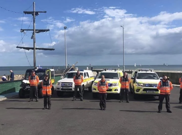 Seven people wearing orange safety vests standing in front of police and emergency vehicles on a seaside promenade with a ship's mast and ocean in the background.