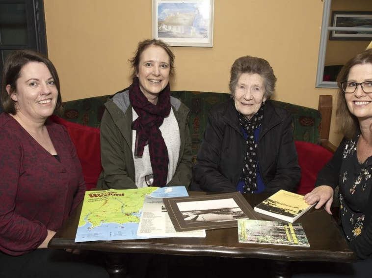 Four women sitting around a table with maps and books, smiling for a photo inside a cafe or restaurant.
