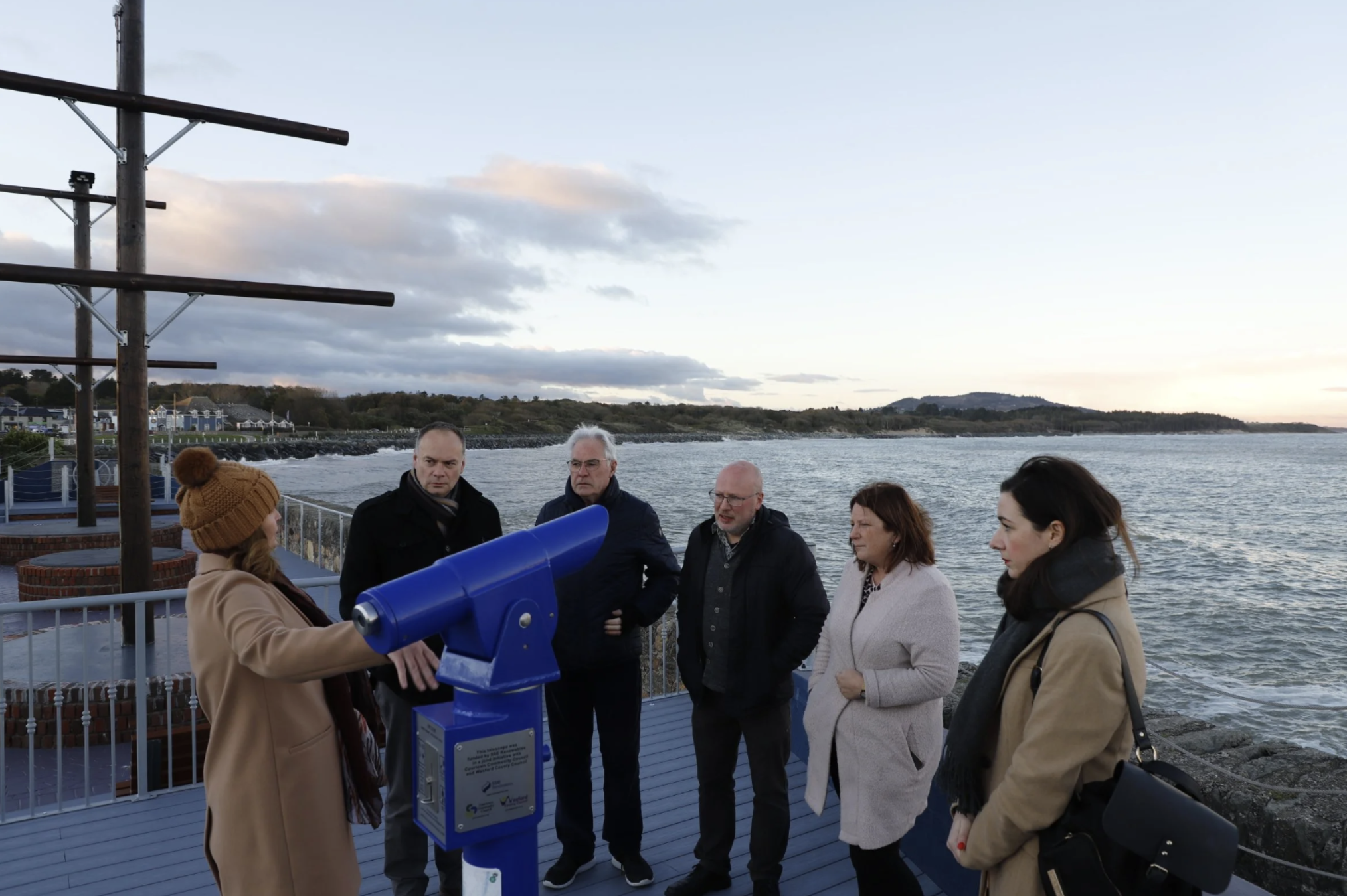 A group of people standing on a coastal outdoor area during the evening, looking through a blue telescope. The background features water, a cloudy sky, and distant land.