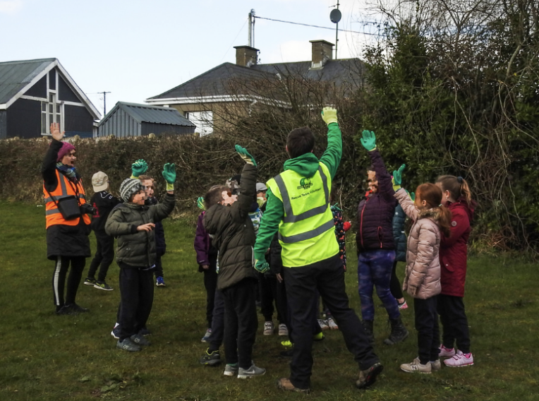 Group of children and adults outdoors during daytime, some raising their hands, with a man in a high-visibility vest leading an activity near bushes and trees.