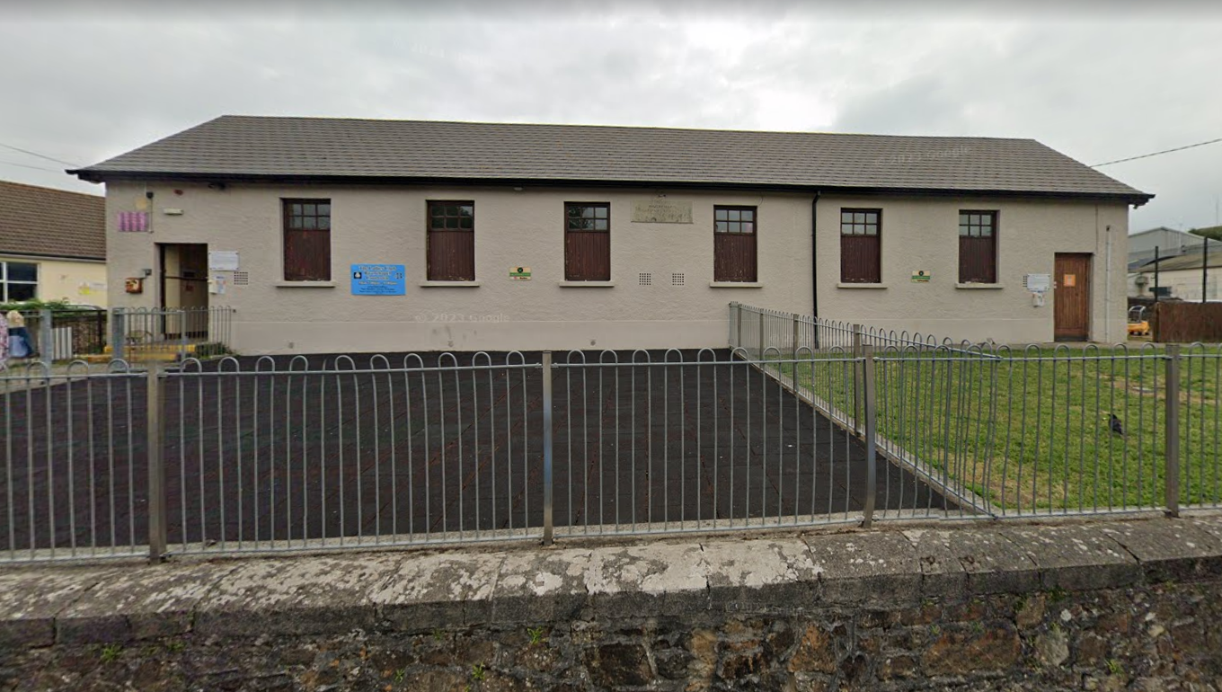 A beige building with a grey roof, multiple windows with dark shutters, and two doors, one on each end. There is a parking area in front, enclosed by a metal fence, and a small grassy area to the right.