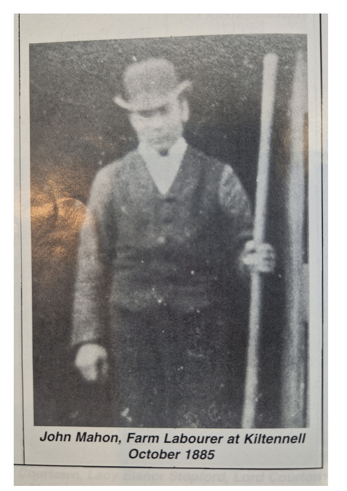 Black and white photograph of a man wearing a bowler hat, suit, and tie, holding a long cane, standing indoors. Caption below reads: "John Mahon, Farm Labourer at Kiltenell October 1885."