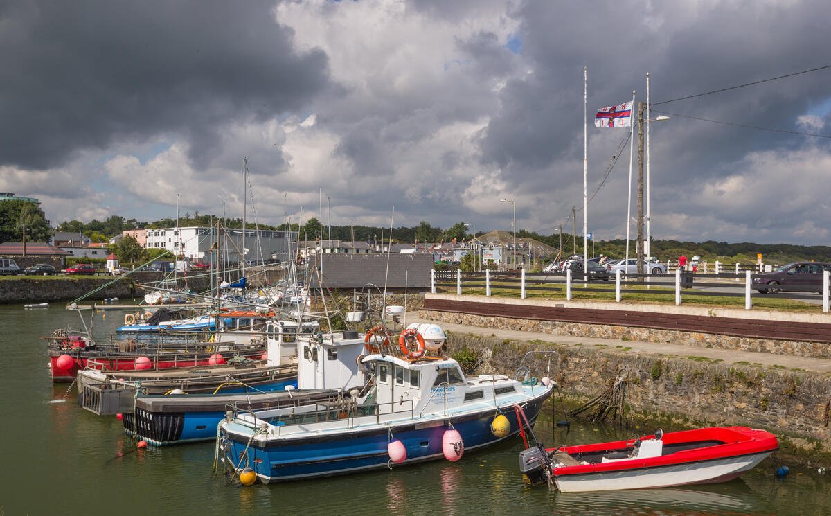 Boats docked along a harbor with a parking lot and buildings in the background, overcast sky with dark clouds.
