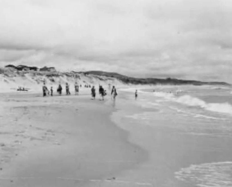 Black and white photo of people walking along a beach near some cliffs under cloudy skies.