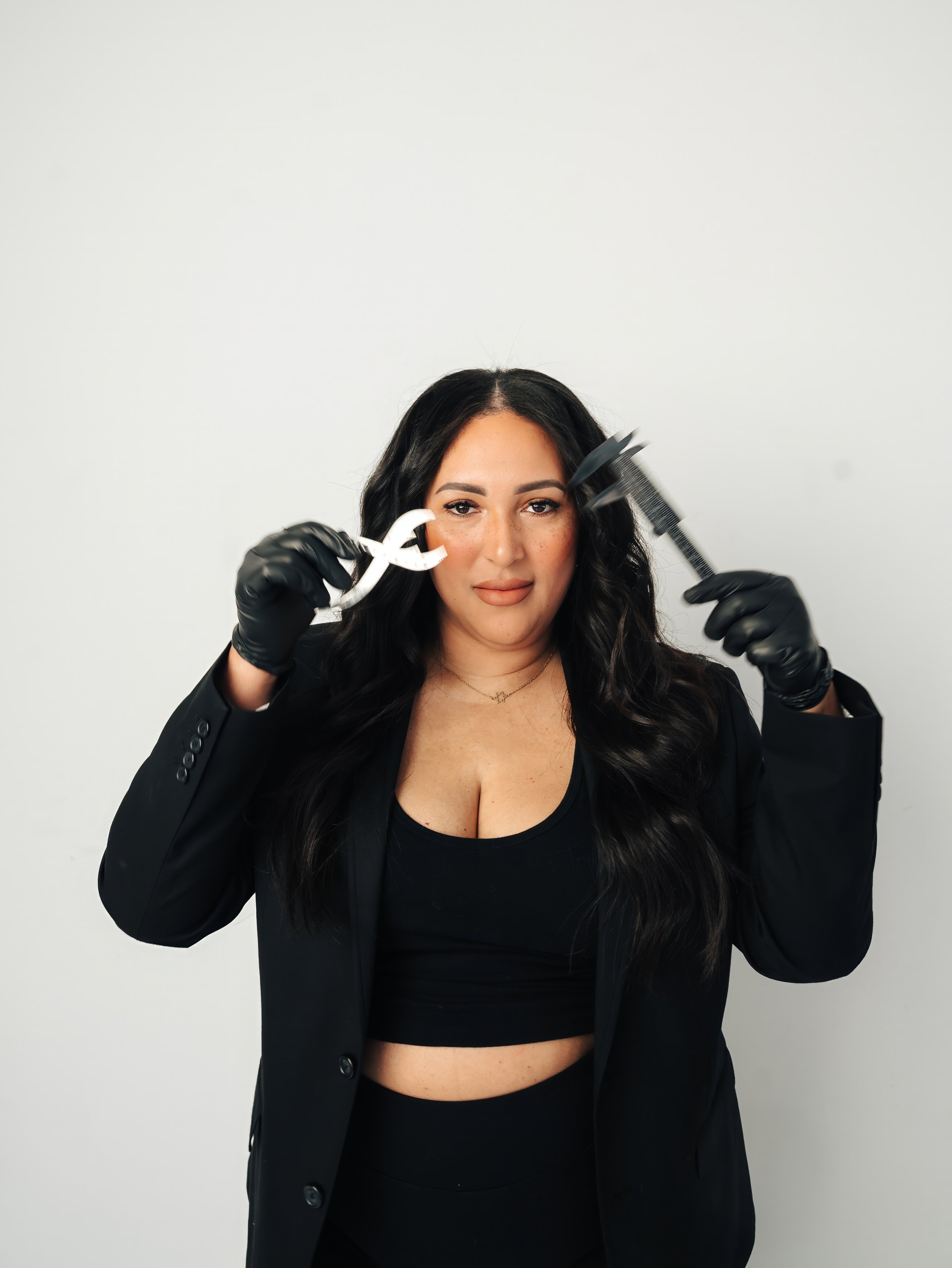 Woman with dark, wavy hair and a black outfit holding a wrench and a pair of pliers, standing against a plain white background.