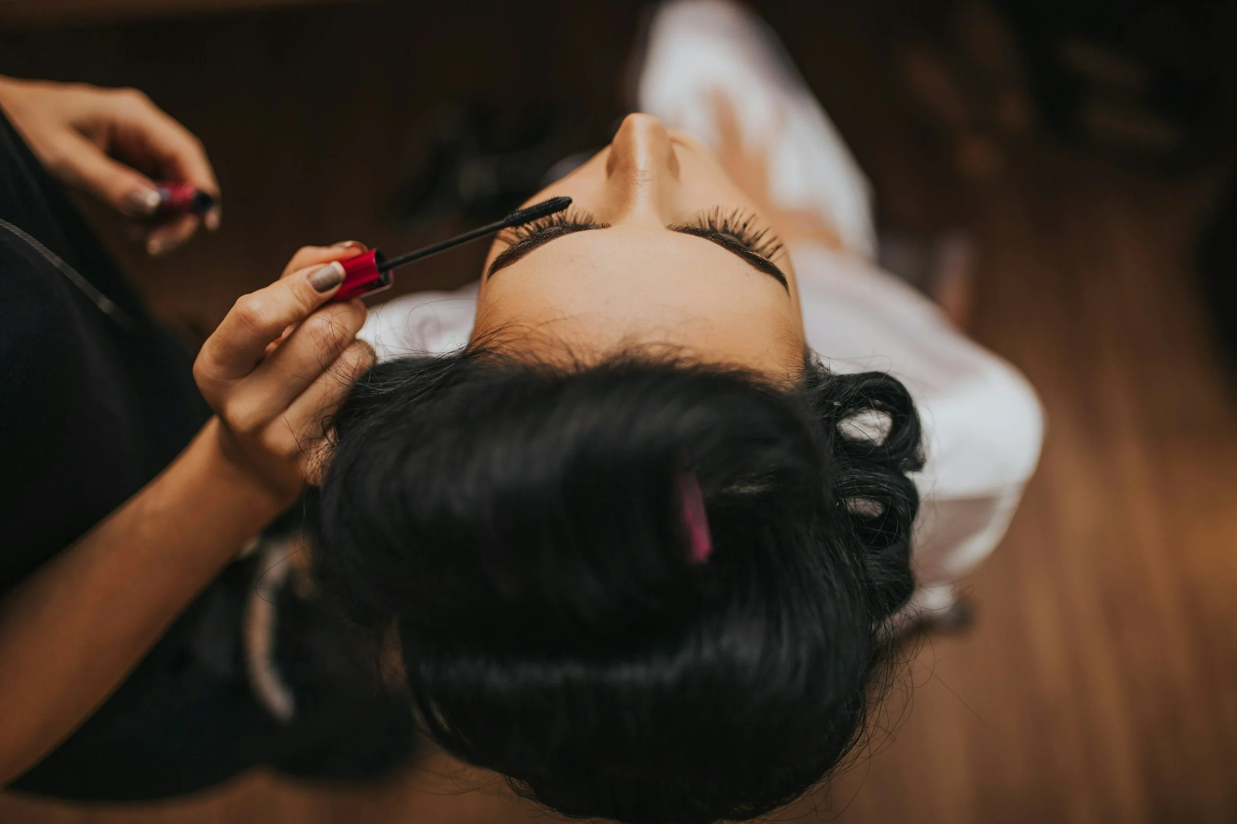 A person with dark hair lying down, applying mascara to their eyelashes with a brush.