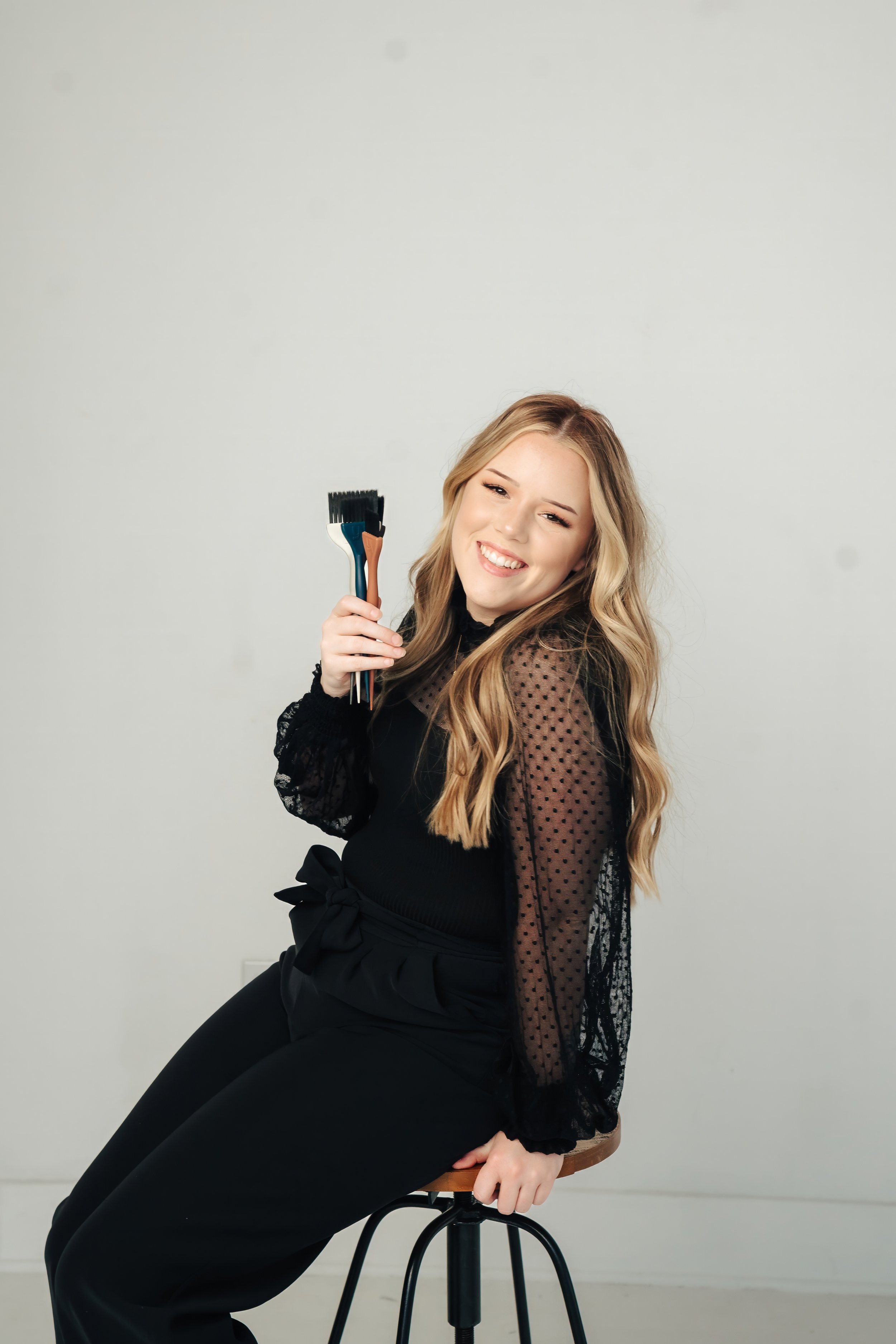 A woman with long blonde hair smiling while sitting on a stool and holding a hair coloring brush. She is wearing a black outfit with sheer sleeves and polka dots, and a bow at her waist.