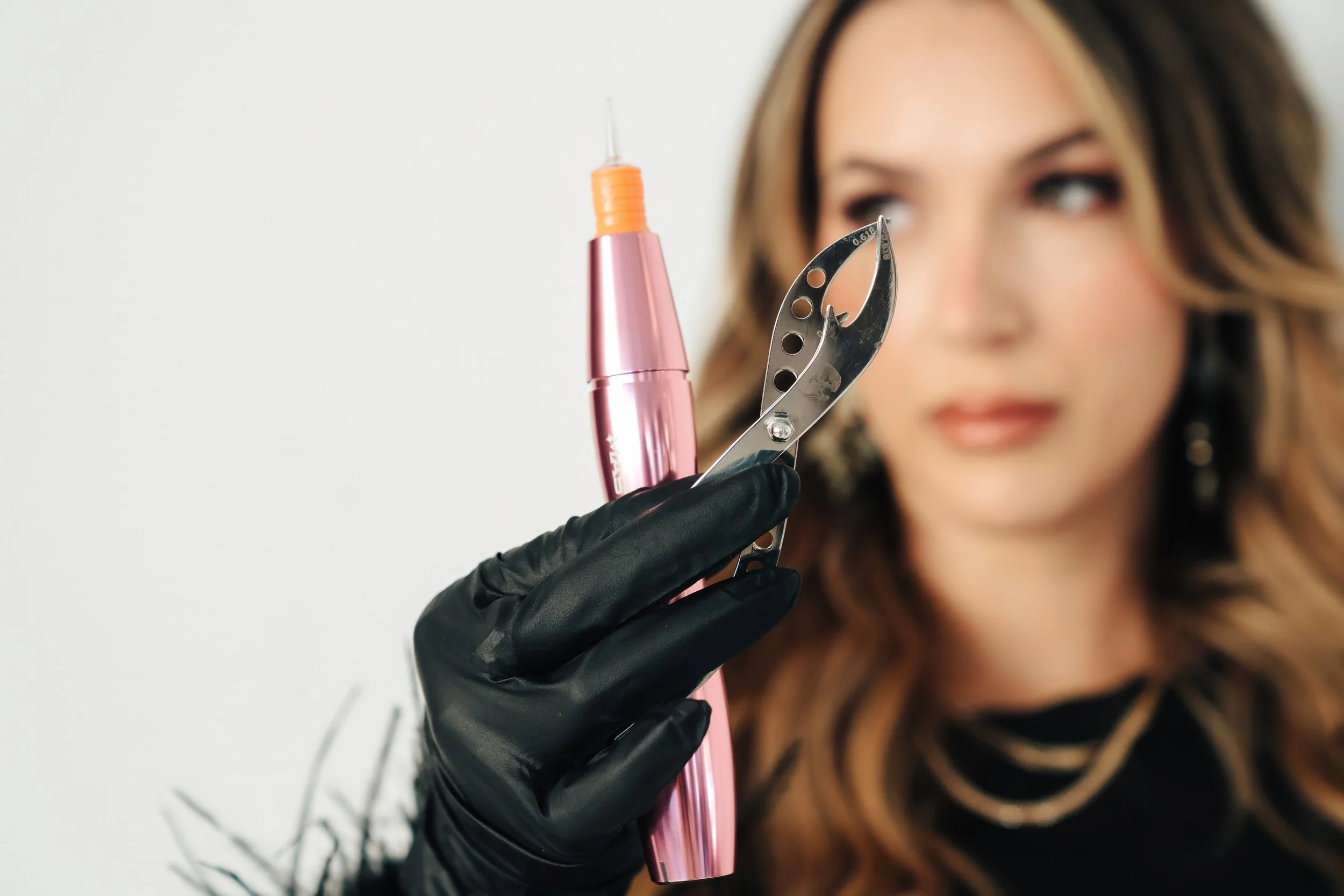 A woman with long, wavy hair, wearing a black glove, holds a pink tattoo machine and a needle, standing against a plain white background.