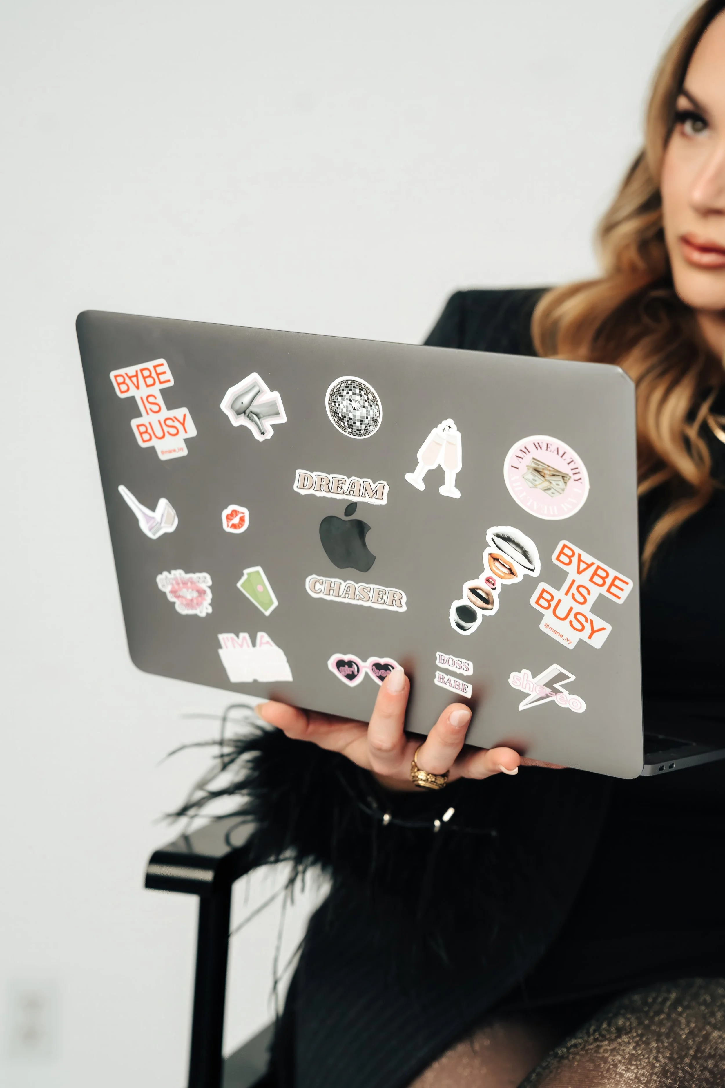 A person holding a silver laptop decorated with various colorful stickers, including comic style speech bubbles, logos, and patterns, with part of their face and hand visible.