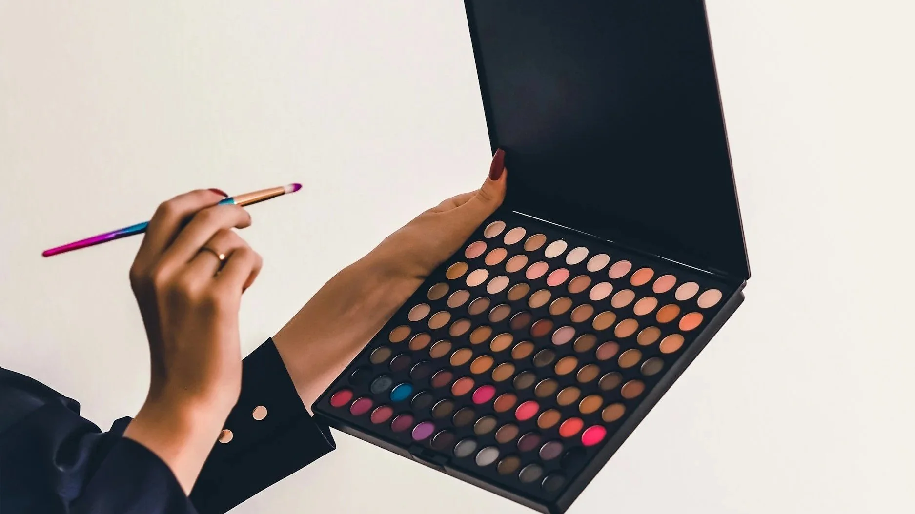 Person holding a colorful makeup brush and open palette of various eyeshadows on a white background.