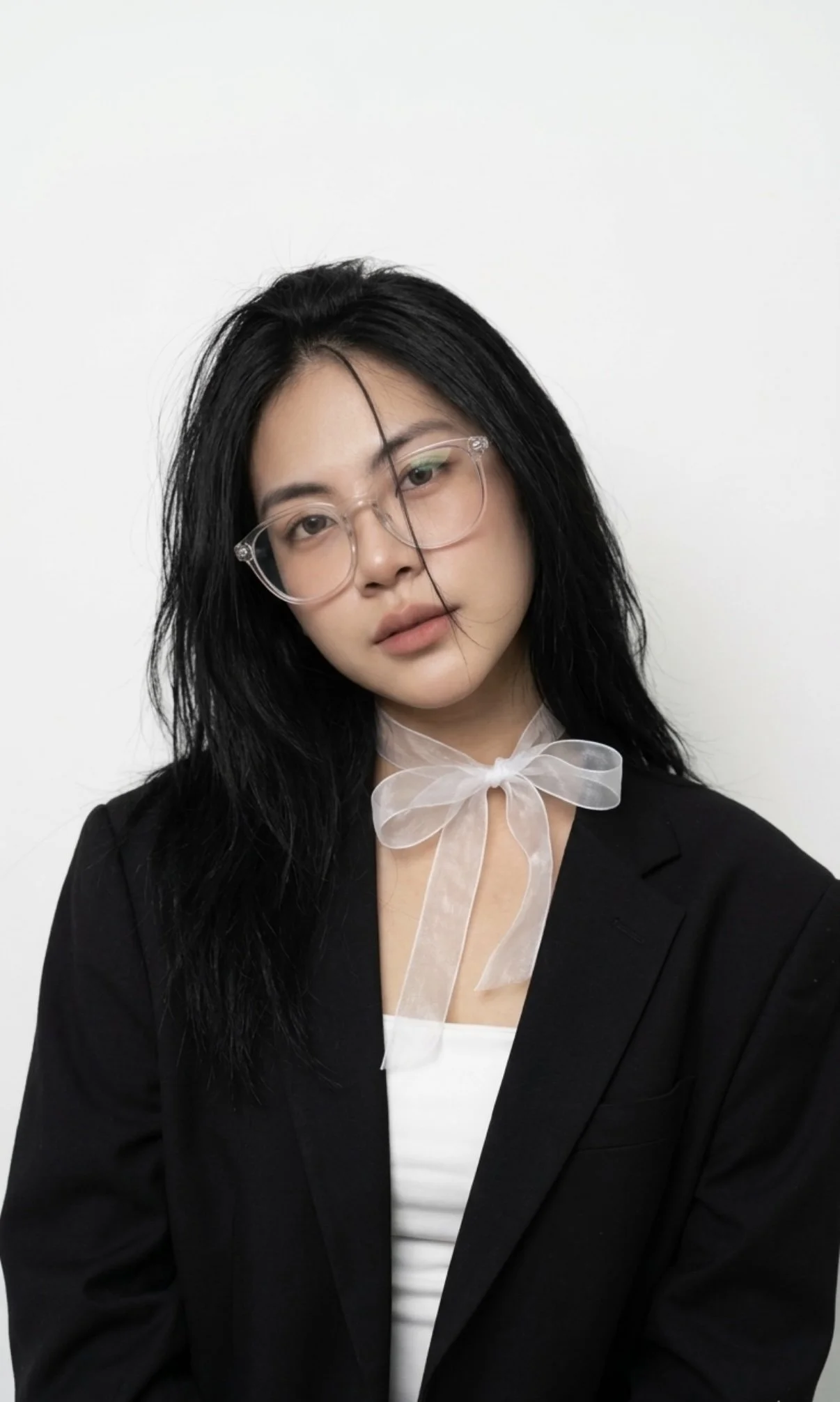 Portrait of a young woman with black, slightly messy hair, wearing large clear glasses, a white top, a black blazer, and a transparent ribbon tied into a bow around her neck, against a plain white background.