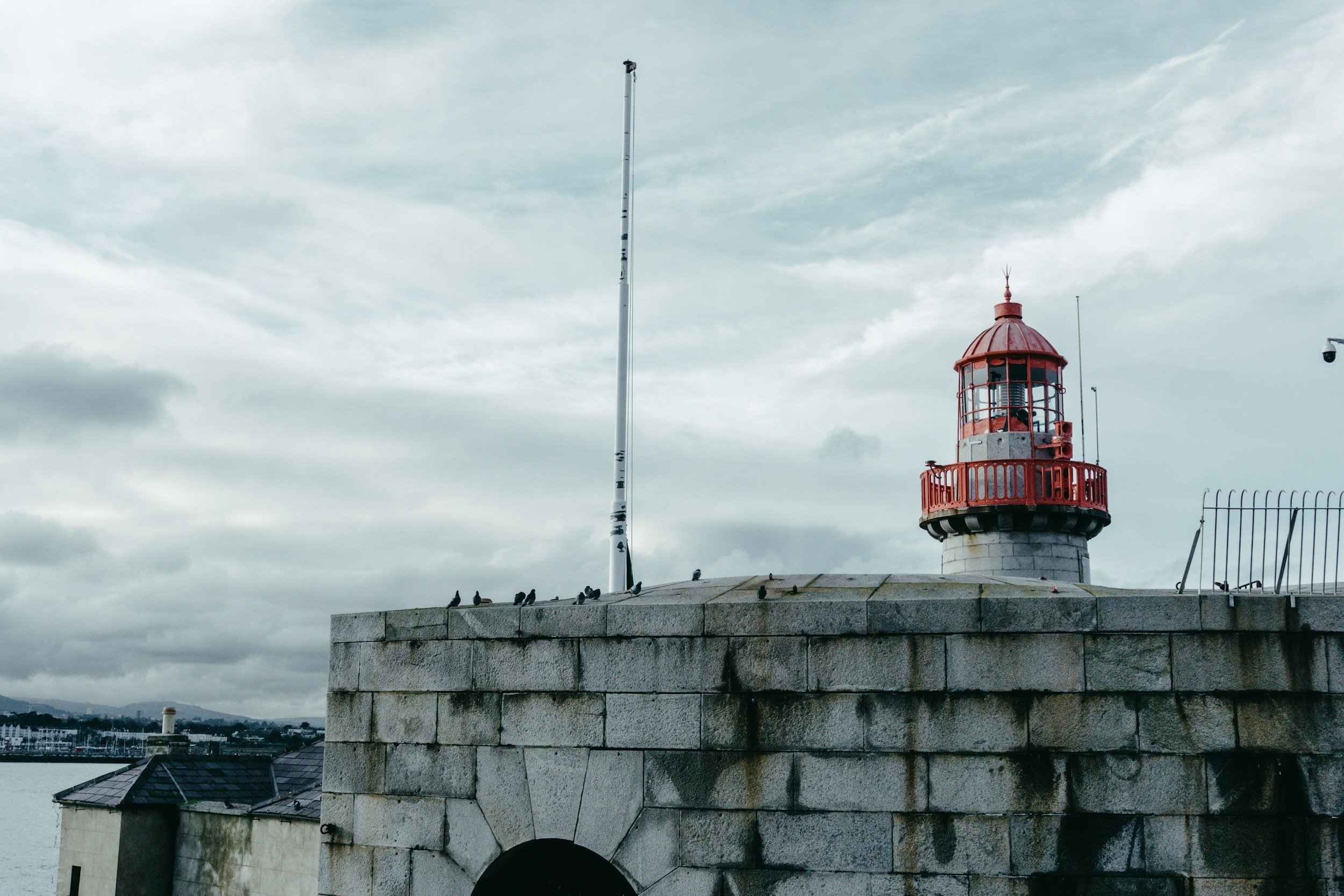 A lighthouse with a red top and railing on a stone platform, with pigeons and a cloudy sky in the background.