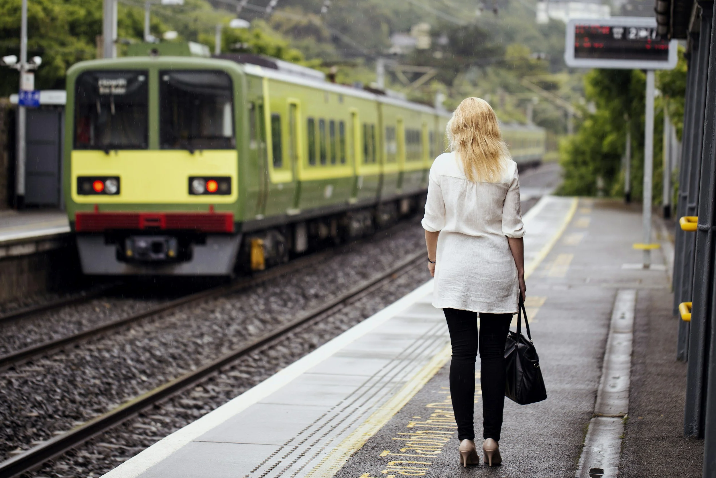 A woman with blonde hair wearing a white jacket and black pants waiting at a train station platform as a green train approaches.