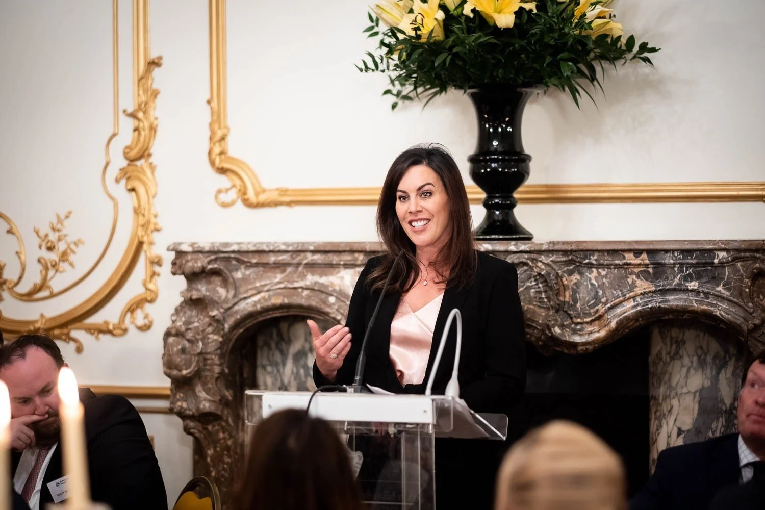 A woman speaking at a podium during a formal event. She has long dark hair and is wearing a black blazer with a light-colored top. There is a marble fireplace and decorative gold trim in the background, and people sitting around her.