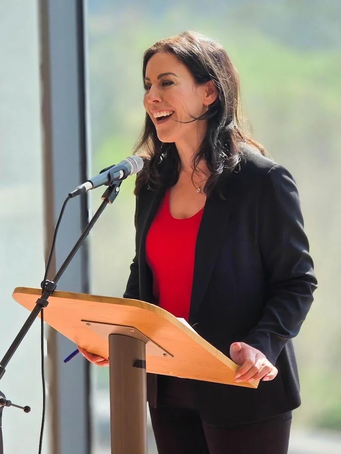 A woman with dark hair, wearing a black blazer and red top, speaking at a podium with a microphone in front of a large window with a blurred green outdoor background.