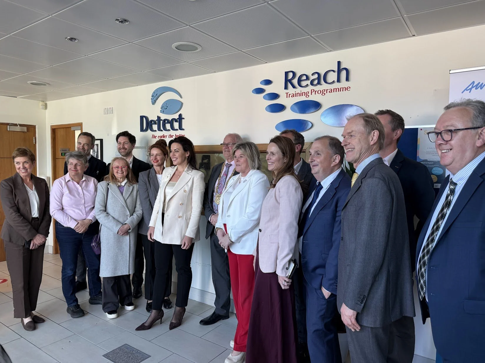 A group of professionals standing together inside a building, smiling for a photo, near a wall with signs that read 'Detect' and 'Reach Training Programme.'