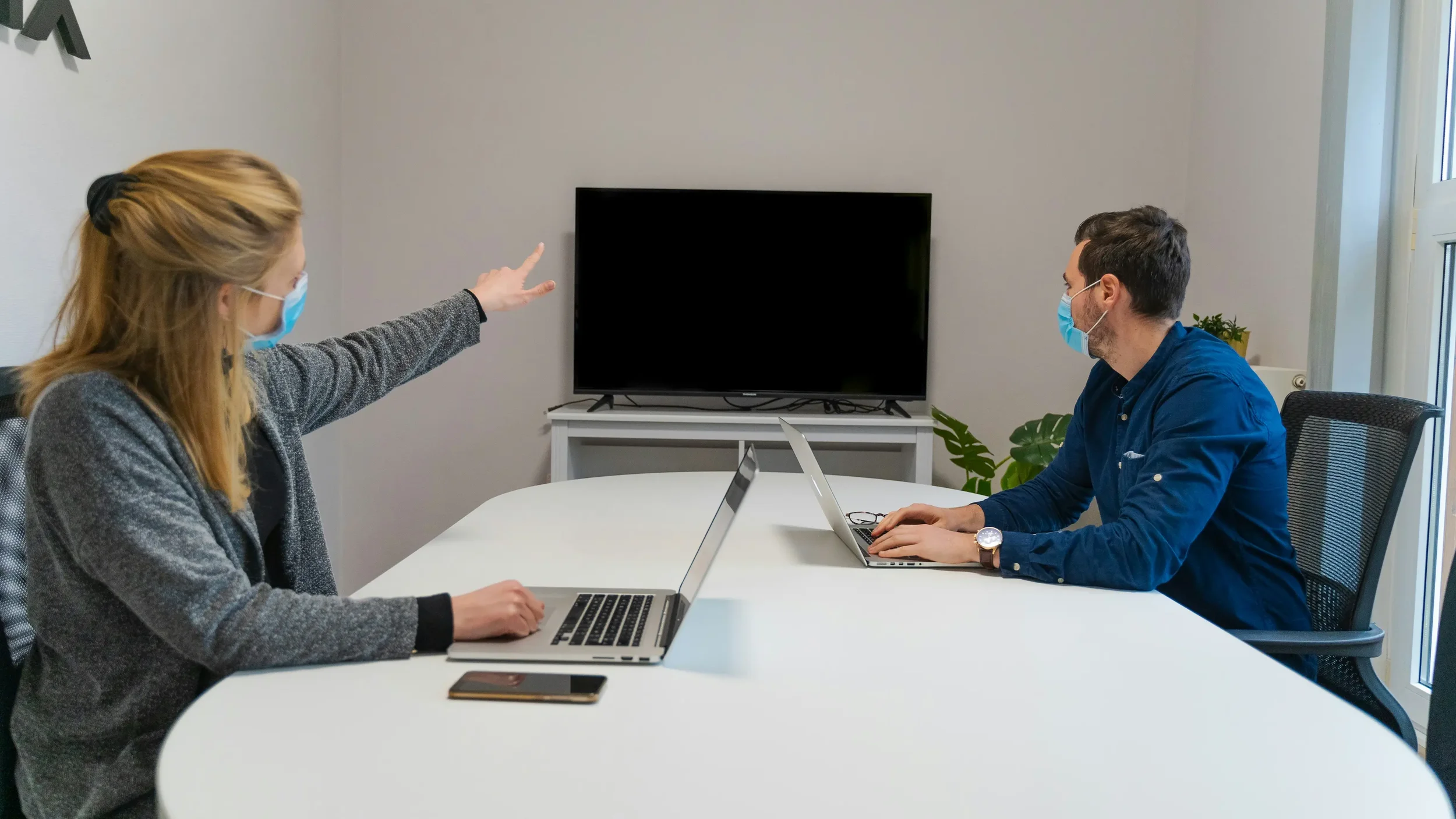 Two people in masks sitting at a conference table with laptops, in a room with a television and a plant.