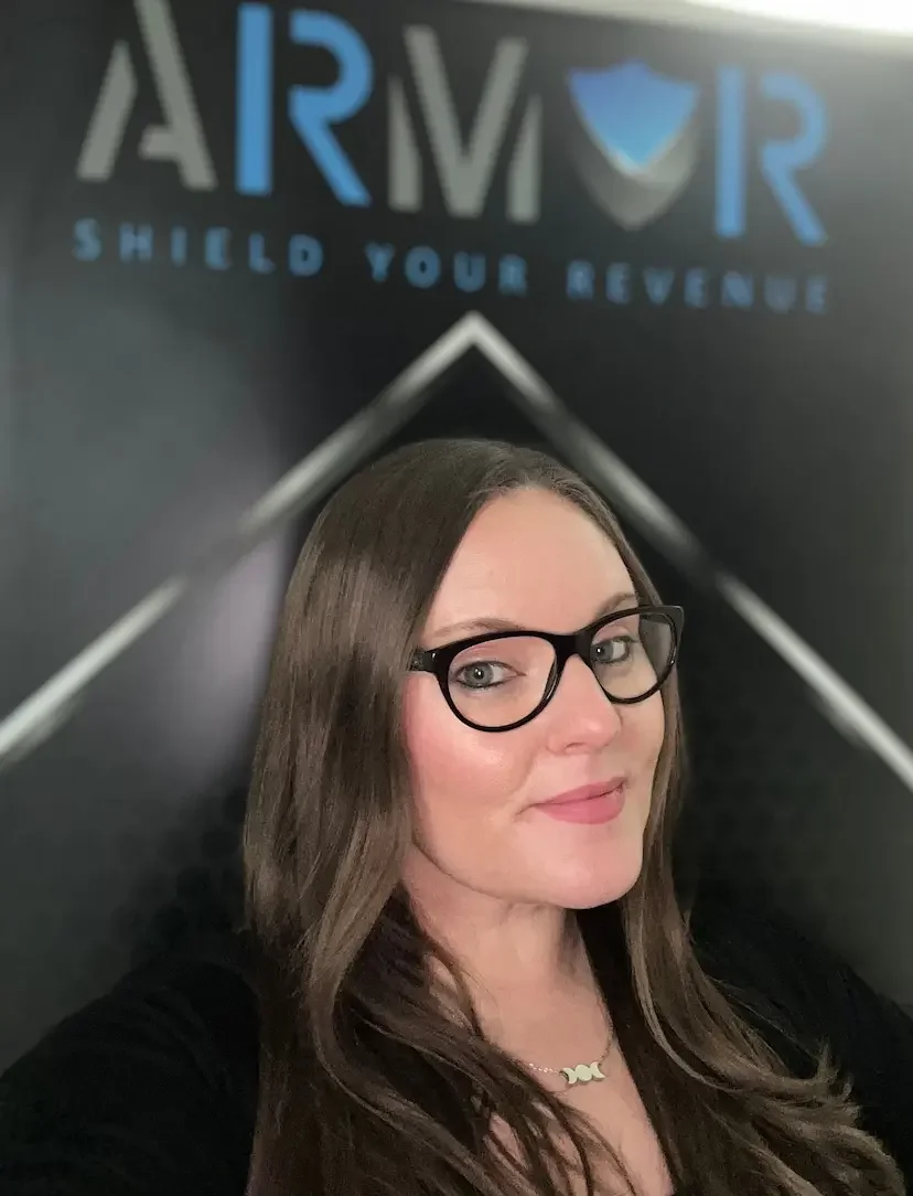 A woman with long brown hair and black glasses (Carmen Frischkorn) taking a selfie in front of a wall with a logo and the words 'Armor Shield Your Revenue' in blue and black.
