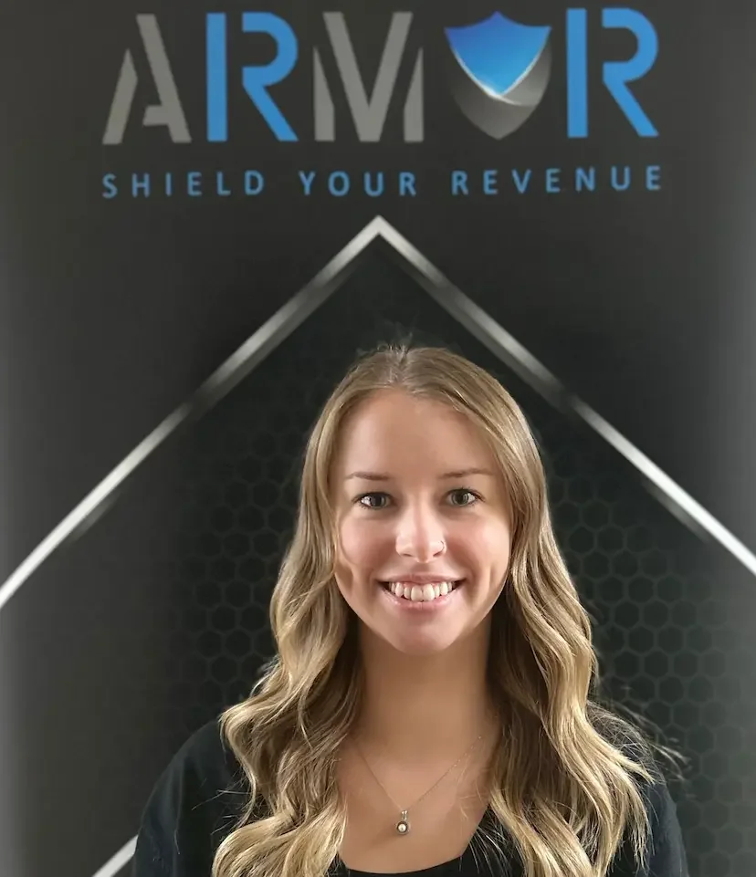 A smiling young woman with long wavy blonde hair (Heather Powell) standing in front of a black background with a logo and the text 'ARMOR' and 'SHIELD YOUR REVENUE'.