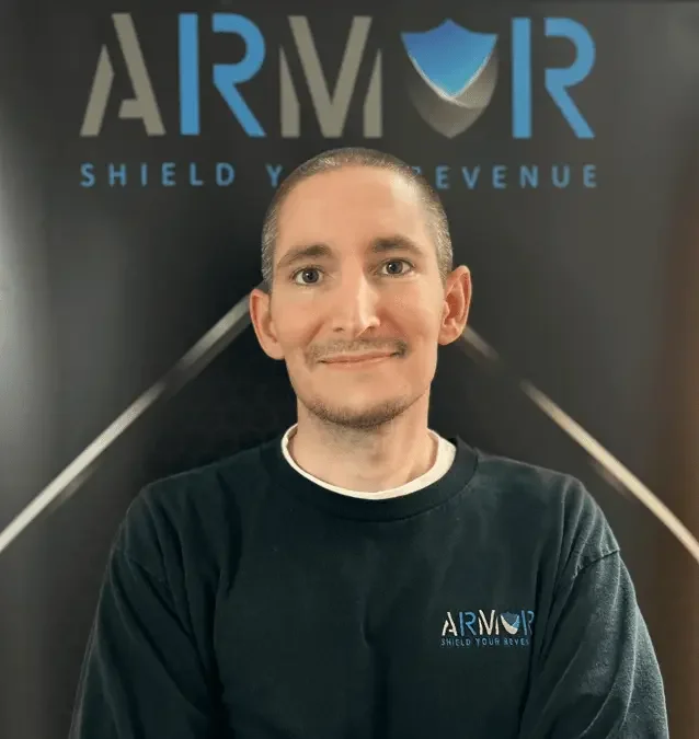 A man wearing a black ARMOR shirt standing in front of a dark background with the ARMOR logo and slogan 'Shield Your Revenue'.