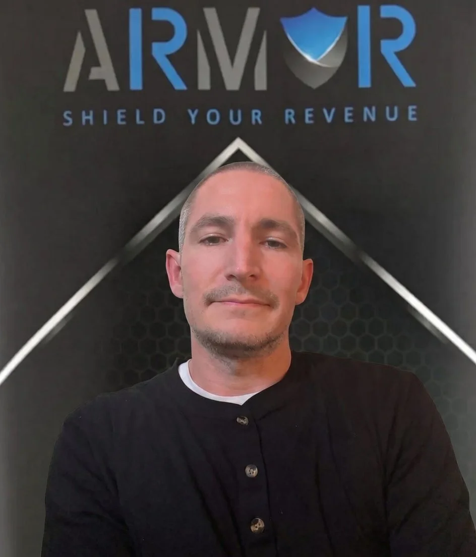 A man (Michael Williams) taking a selfie in front of a black backdrop with the word ARMOR and a shield icon, with the slogan "Shield Your Revenue" underneath.
