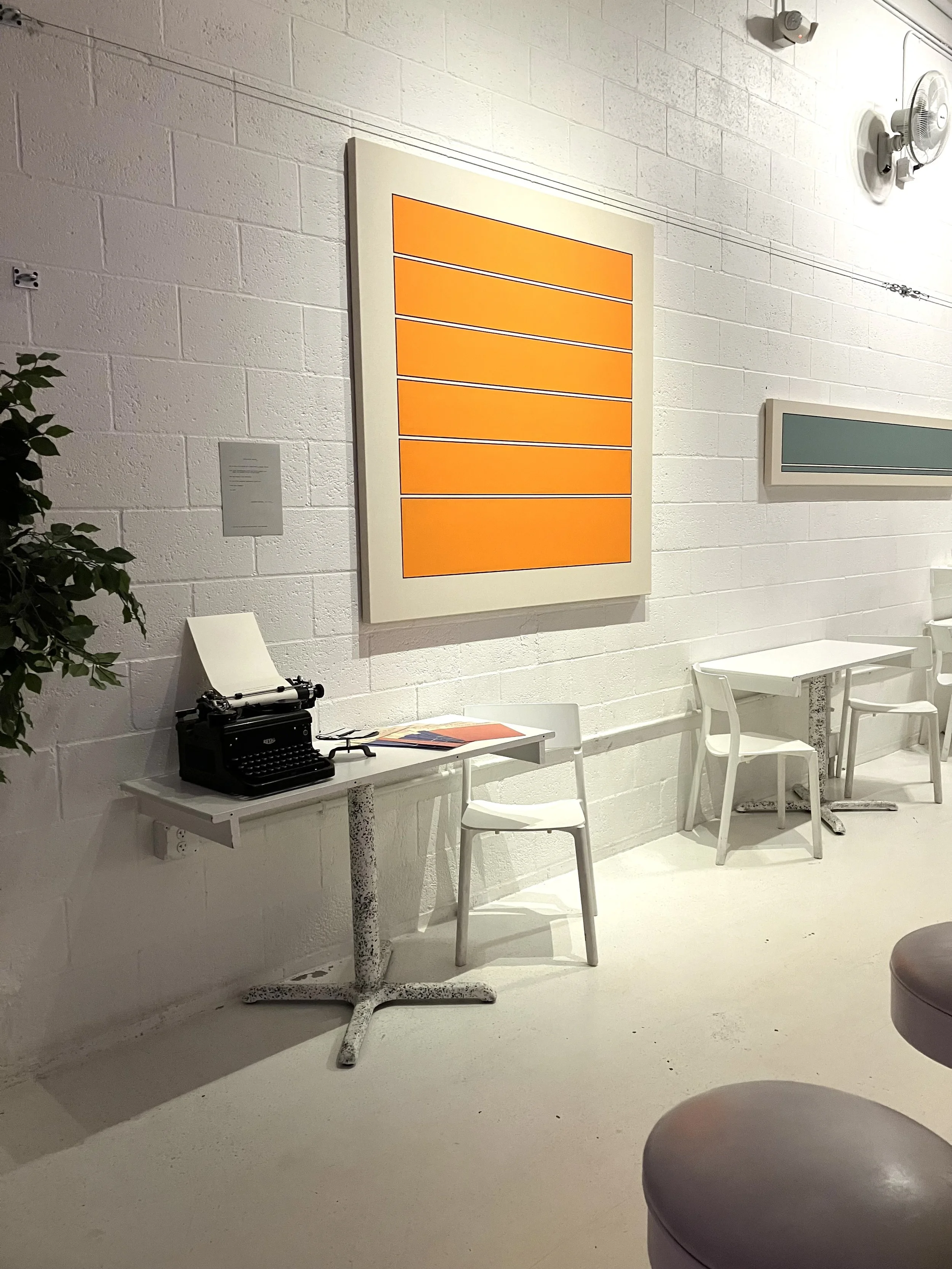 Interior of art gallery imaginary factory with white brick walls, an orange and yellow geometric painting: "Polysocial Colors: Imaginary Orange ", a small white table with a typewriter and some papers, white chairs, a ottoman, a potted plant.