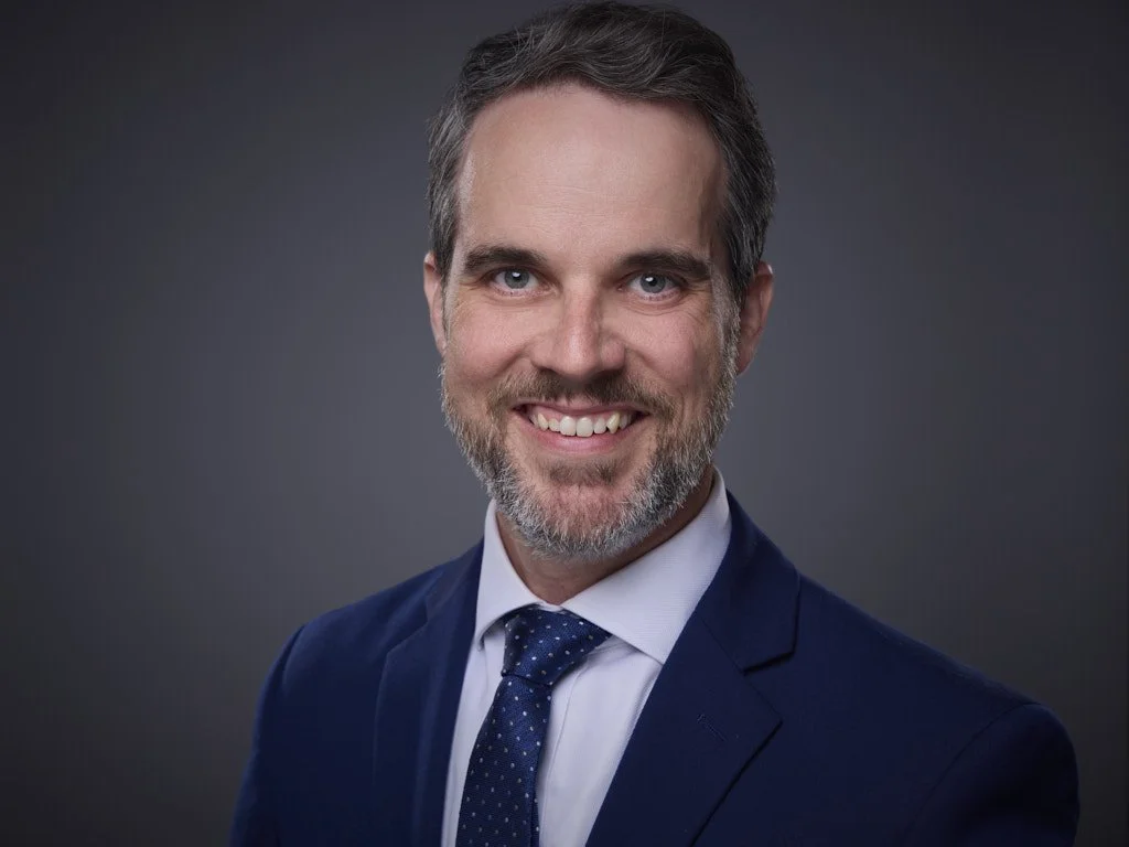 A man with short dark hair and a beard, wearing a navy suit, white shirt, and patterned tie, smiling in front of a dark gradient background.