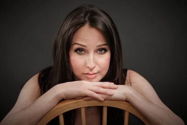 Portrait of a young woman with dark hair resting her chin on a wooden chair back, looking at the camera against a dark background.