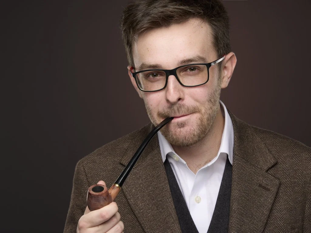 A young man with glasses, beard, and mustache, wearing a white shirt and brown blazer, holding a smoking pipe with a confident look.