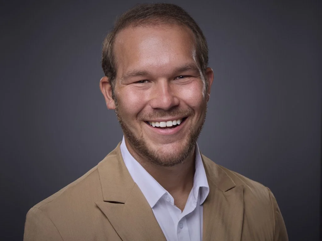 Headshot of a smiling man with light brown hair, a beard, and a mustache, wearing a tan blazer and white shirt, against a dark gray background.