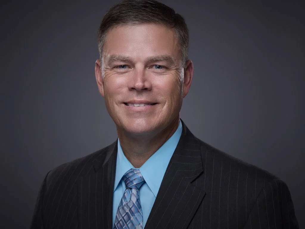 Headshot of a smiling middle-aged man in a business suit and tie against a dark gray background.
