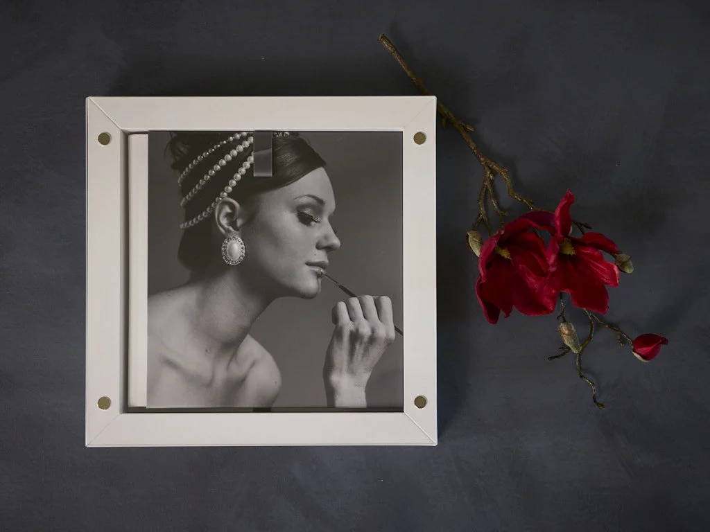 A framed black and white photograph of a woman with vintage hairstyle, pearl earrings, and large earrings, holding a makeup brush near her lips. Next to the frame is a branch with red flowers on a dark surface.