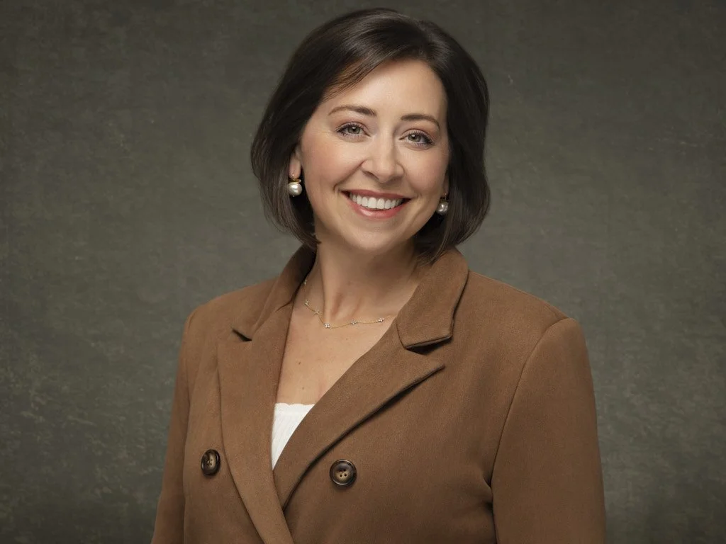 Professional portrait of a woman with short brown hair, smiling, wearing pearl earrings, a brown blazer, and a white top, against a neutral background.