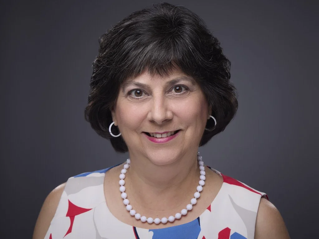 Professional headshot of a smiling middle-aged woman with short dark hair, wearing a sleeveless dress with red, white, and blue patterns, pearl necklace, and hoop earrings, against a dark gray background.