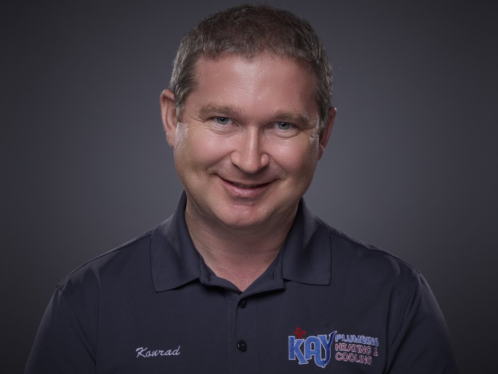 Professional headshot of a smiling man with short brown hair wearing a black polo shirt with an embroidered logo for Kay Plumbing Heating & Cooling and a name tag reading 'Kawrad' against a dark gray background.