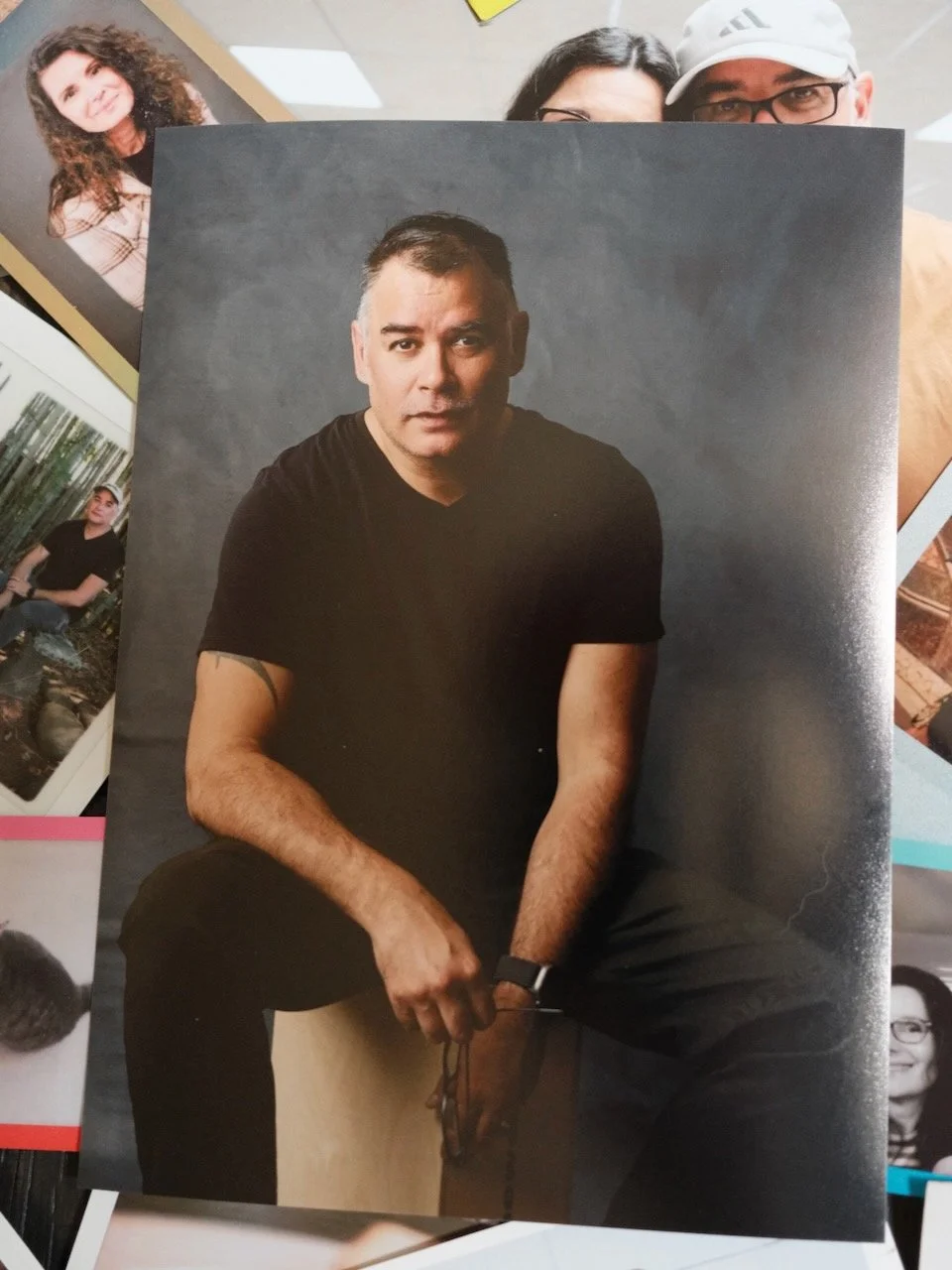 A man with short dark hair wearing a black t-shirt, sitting with a serious expression against a dark background.