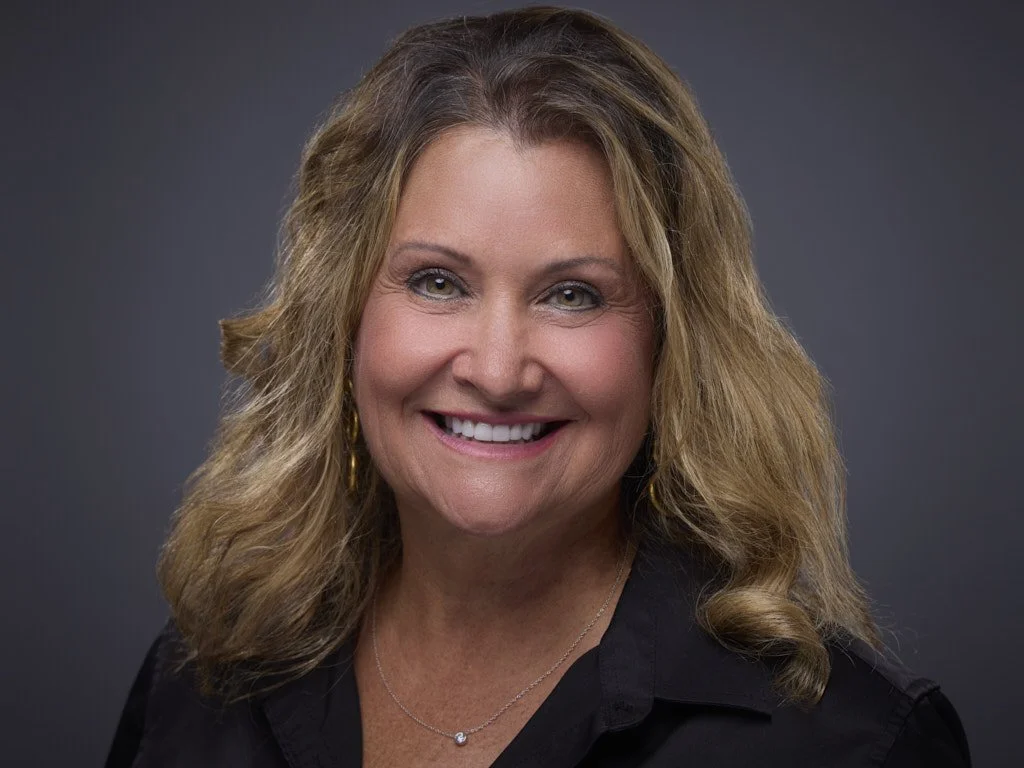 Headshot of a woman with blonde, wavy hair, smiling, wearing a black top and a necklace, against a gray background.