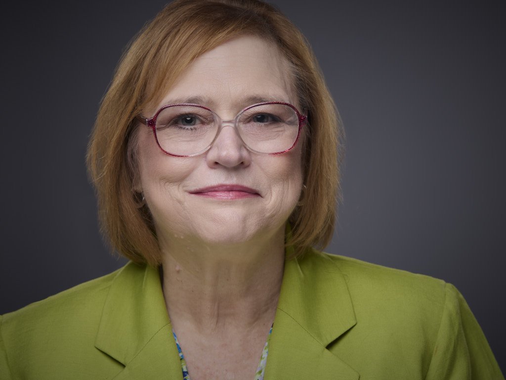 Professional headshot of a woman with shoulder-length red hair, wearing glasses with red frames, a green blazer, and a multicolored top, smiling gently against a dark gray background.